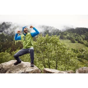 A man wearing a green and blue knitted fleece vest poses on a rock outdoors, surrounded by greenery and mountains.