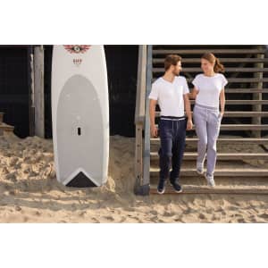 A pair of ladies jog-pants in grey, featured alongside a man and woman on a sandy beach near steps and a surfboard.