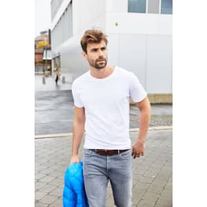 A man in a white T-shirt and grey jeans carries a blue jacket, walking on a paved area near modern buildings.