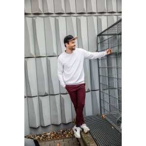 A man is wearing a white jumper and burgundy pants, standing next to a wire fence. He has a black cap on.