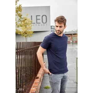 A navy blue T-shirt worn by a man standing outdoors near a fence and a building in a urban setting.