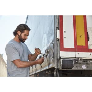 A man in a grey T-shirt uses a wrench on a truck's latch. The setting appears industrial or outdoor.