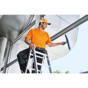 A man in an orange work T-shirt and cap is cleaning windows while standing on a ladder. The setting features modern architecture.