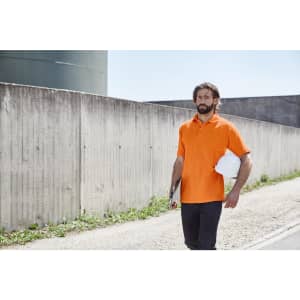 An orange work polo shirt on a man walking outdoors, holding a white helmet and a clipboard.