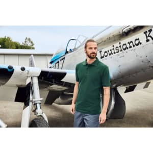 A man stands beside a vintage aircraft, wearing a dark green work polo shirt and grey trousers. The aircraft features a logo.
