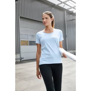 A light blue women's work t-shirt is displayed on a model with black pants, set in an industrial environment.
