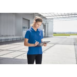 A woman in a blue work polo shirt stands outdoors, holding a tablet and taking notes. The setting features a large, open space.