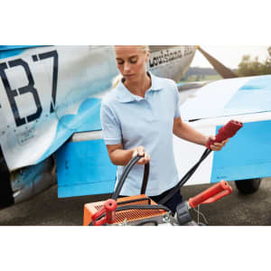 A woman in a light blue polo shirt holds equipment near an aircraft, showcasing a professional workwear style.