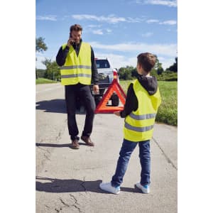 Two individuals wearing bright yellow safety vests stand by the roadside. The adult is on a phone, while the child holds a reflective triangle.