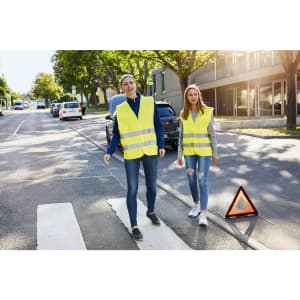 Two adults wear bright yellow safety vests while walking on a street, with a warning triangle nearby.
