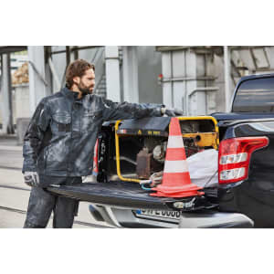 A man in a dark grey workwear jacket stands by a pickup truck, examining equipment inside the vehicle's tray.