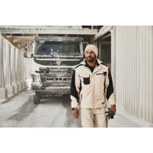 A man wearing a white and black Craftsmen softshell jacket stands in a construction setting, with a truck in the background.