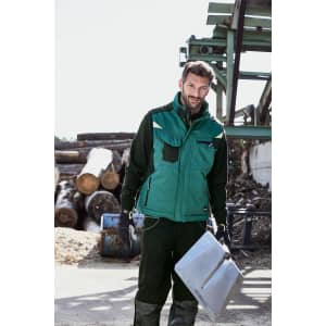 A man stands outdoors wearing a green softshell vest over a black outfit, holding a metal sheet. Logs and machinery are in the background.