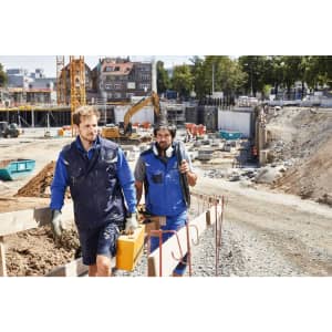 Two men wearing Craftsmen Softshell Vests in blue walk on a construction site with tools in hand.