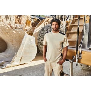 A man stands in a beige Craftsmen T-Shirt, beside heavy machinery in a rugged outdoor setting.