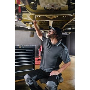 A man in a grey polo shirt works under a vehicle, using a wrench in a workshop setting with tools in the background.