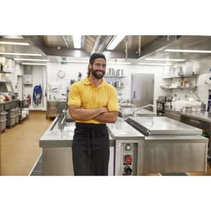 A man in a yellow polo shirt and black apron stands in a clean, modern kitchen, smiling confidently.