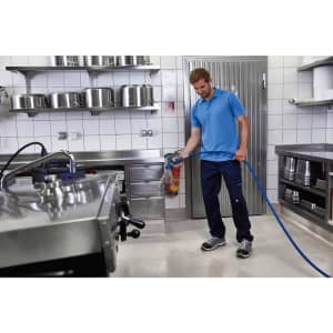 A man in a blue polo shirt and navy trousers stands in a commercial kitchen, holding a hose near a work surface.