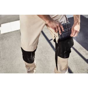 Light beige workwear pants with black knee patches, featuring a pocket being accessed. There is a logo present.