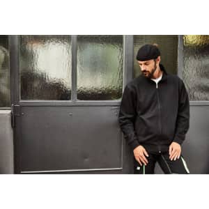 A black workwear sweat jacket worn by a man leaning against a grey door, featuring a logo on the chest.