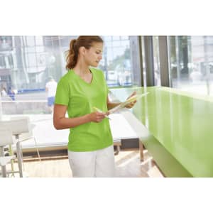A green ladies workwear t-shirt displayed on a woman standing indoors, holding a clipboard. The setting features natural lighting.