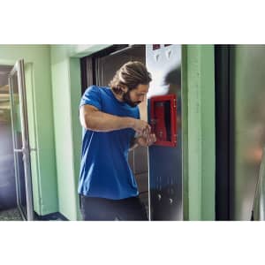 A man in a blue workwear t-shirt adjusts equipment in a corridor, with green walls and metallic surfaces around him.