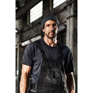 A men's workwear T-shirt in black, featuring a plain design, worn under black overalls in an industrial setting.