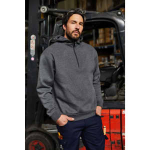 A man in a grey half zip hoodie stands confidently in a warehouse setting with a forklift in the background.