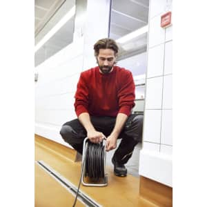A man in a red sweatshirt kneels while managing a hose reel in a tiled hallway. He wears black pants and shoes.