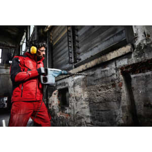 A man in a red workwear softshell jacket is using a power drill in an industrial setting. He is wearing ear protection.