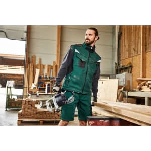 A man wearing a green softshell work vest stands in a workshop, holding a power tool, surrounded by wooden materials.