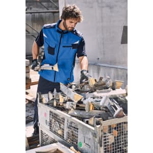 A man in a blue workwear softshell vest inspects metal tools at a construction site. The vest features pockets and a logo.