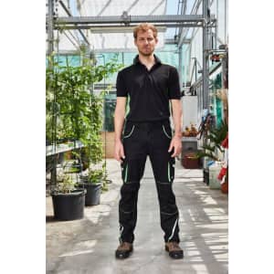 Workwear pants in black with green accents on a model, set in a plant-filled greenhouse. The model wears a black polo shirt.