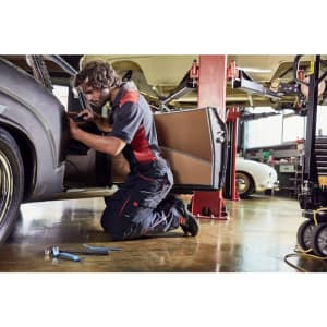 Workwear pants in black with red accents, worn by a person kneeling beside a car in a workshop setting.