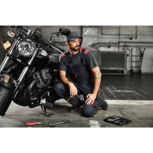 A man kneels beside a motorcycle, wearing blue workwear pants with a bib and tools nearby, set in an industrial environment.