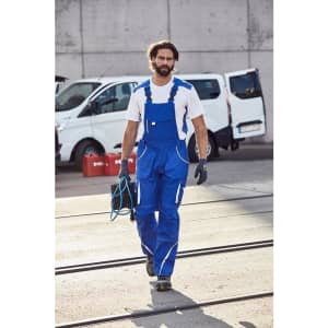 A person wearing blue workwear pants with bib and gloves, holding cables, walking on a construction site.
