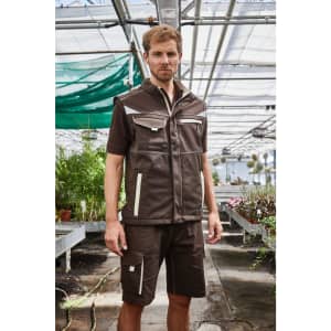 A man models a brown softshell vest with pockets in a greenhouse setting, surrounded by plants.