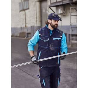 A worker wearing a navy blue softshell padded vest with turquoise accents, holding a metal rod in an outdoor setting.