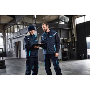 A padded work vest in navy blue with teal accents, worn by two people in a workshop setting, one with a clipboard.