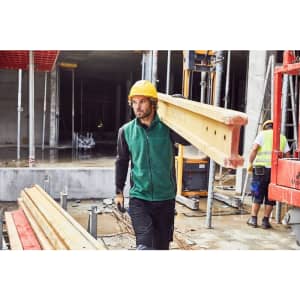 A man in a green fleece vest carries wooden beams at a construction site, wearing a hard hat and work trousers.