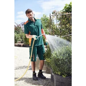 A man in a green and orange work polo shirt waters plants with a hose in a garden setting.