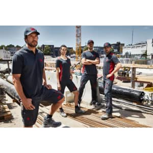 Four individuals wearing black and red men's workwear polos stand on a construction site surrounded by building materials.