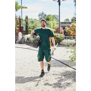 A model wearing a green work t-shirt and shorts, carrying a crate of plants in a garden setting.