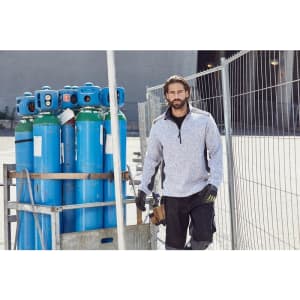 A man wearing a grey knitted fleece half-zip top stands near blue gas cylinders, with a worksite backdrop.