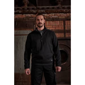 A man wearing a black knitted workwear fleece half-zip, set against a rustic industrial background.