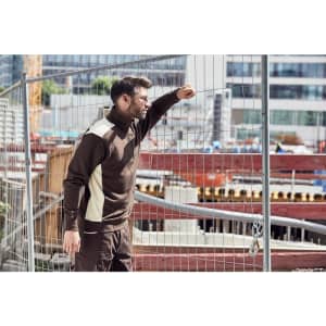 A man stands beside a construction site wearing a brown and cream half-zip work jumper with a logo.