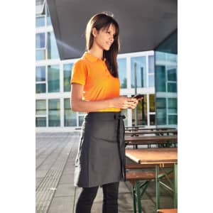 A woman wearing an orange polo shirt and a grey skirt stands outside, holding a mobile phone.