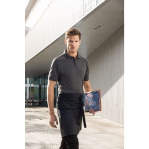 A man in a grey polo shirt and apron holds a menu while walking outside a modern building.