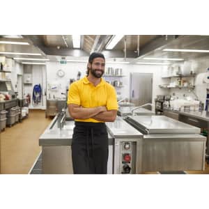 A man in a yellow polo shirt stands confidently in a commercial kitchen, featuring stainless steel equipment and a tidy environment.