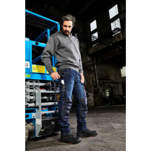 A man wearing grey workwear and blue stretch jeans stands near a worksite with industrial equipment.